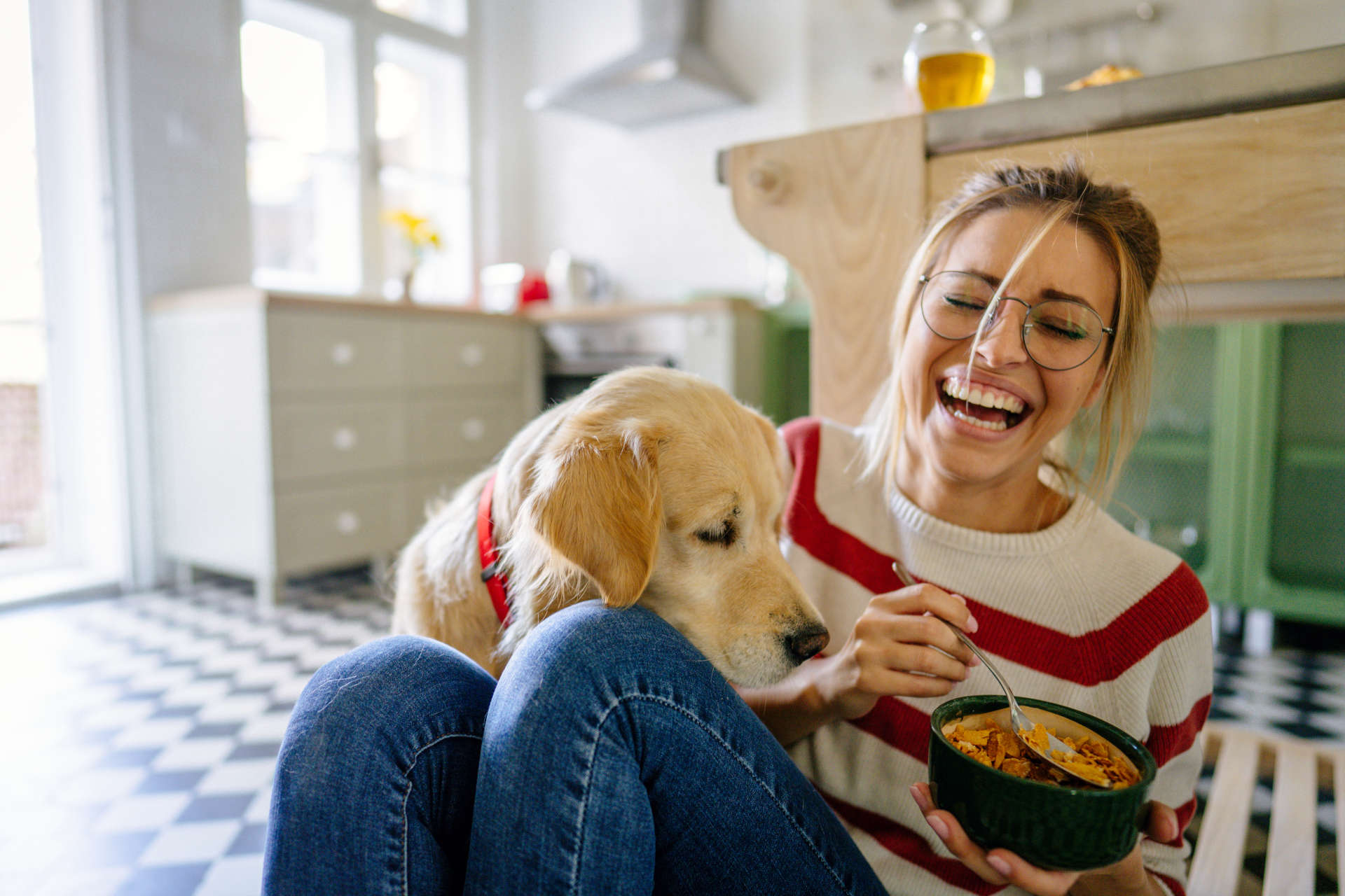 A young person sitting on a kitchen floor who is laughing while they try to prevent a yellow Labrador Retriever dog from eating the food from the spoon and bowl that they're holding.