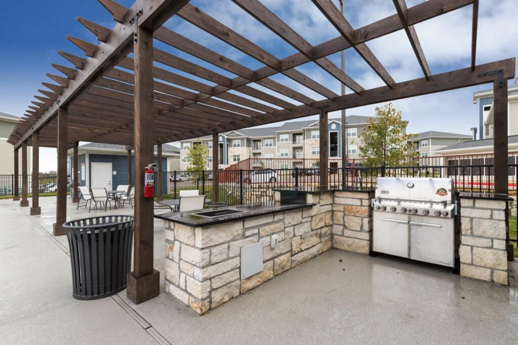 Exterior grilling station with a wooden pergola, stainless steel grill, counter tops and prep areas, two sets of patio tables with chairs, a trash can, and apartment resident and community buildings in the background.