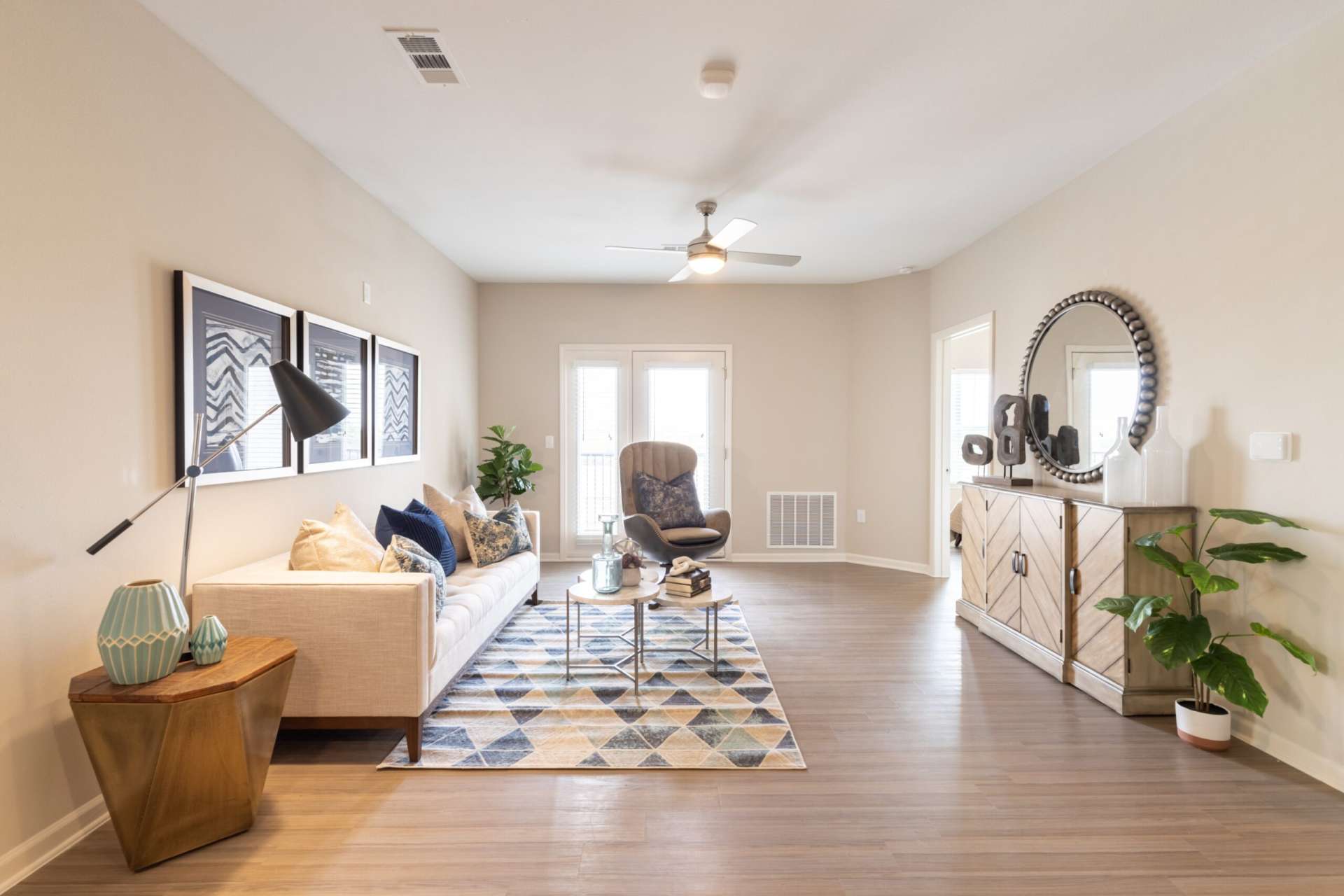 A living room at Copper Pointe apartments with couch, area rug, recliner, end table with lamp, mirror, plants, decor, a cabinet, and plenty of natural light.