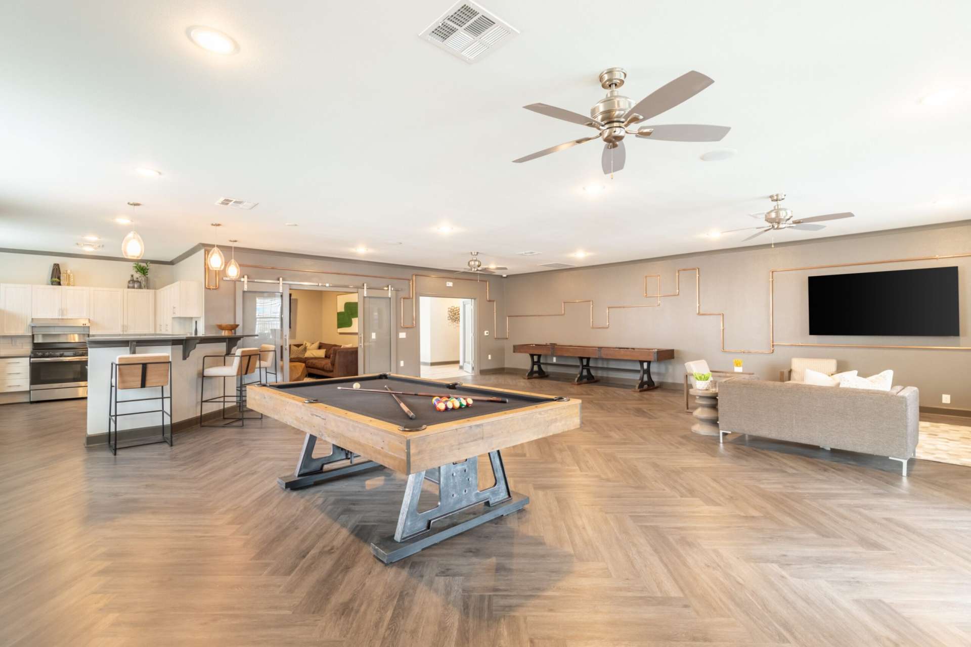 A wide view of the community room with couch, large screen TV, ceiling fans, pool table, game table, countertop with chairs, kitchen area with oven, an a separate lounge space with additional couch and chairs.