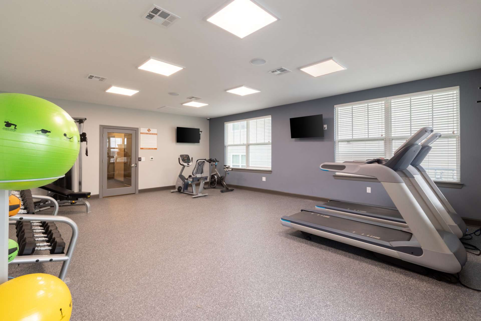 A view of the community fitness center with two treadmills, two stationary bikes, a weightlifting bench, a rack of free weights, a rack of yoga balls, and two large screen TVs mounted on the walls.