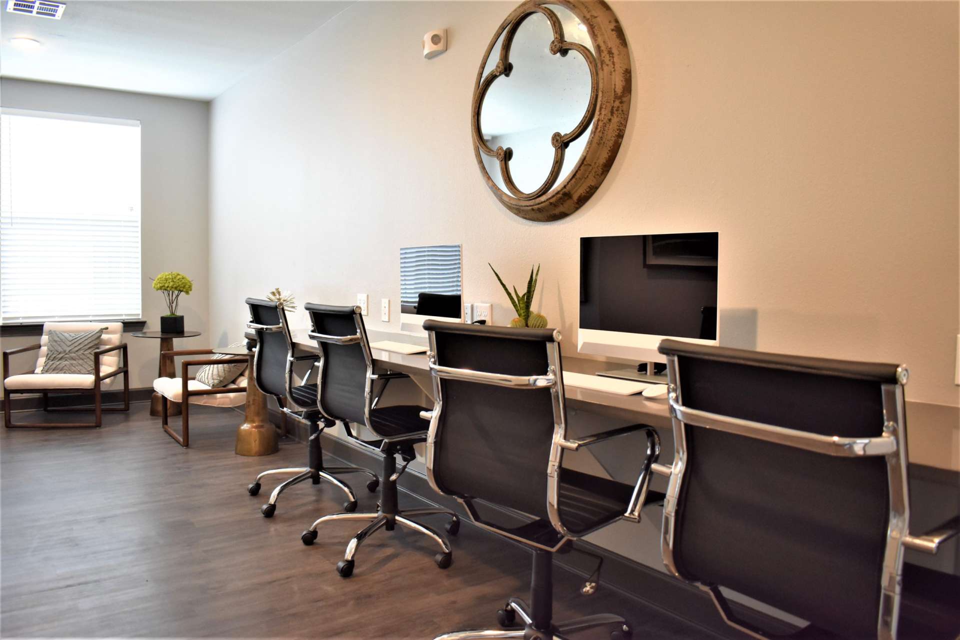 A work station countertop with two computer monitors and keyboards, four desk chairs, a mirror on the wall, and a small table with two lounge chairs.