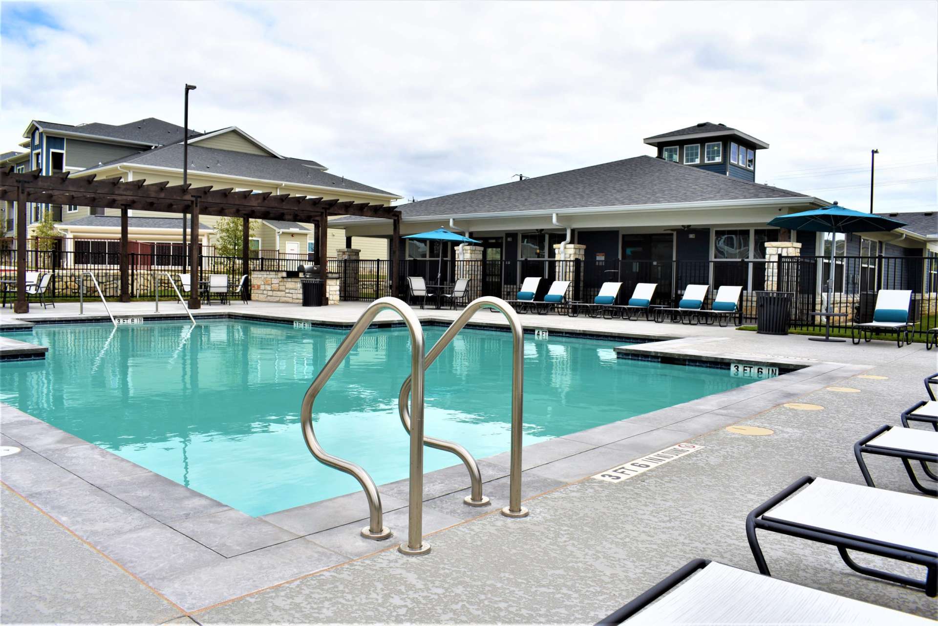 Swimming pool with in-pool ladder and handles, a wrought iron fence surround it, lounge chairs along the deck area, and an apartment building in the background.