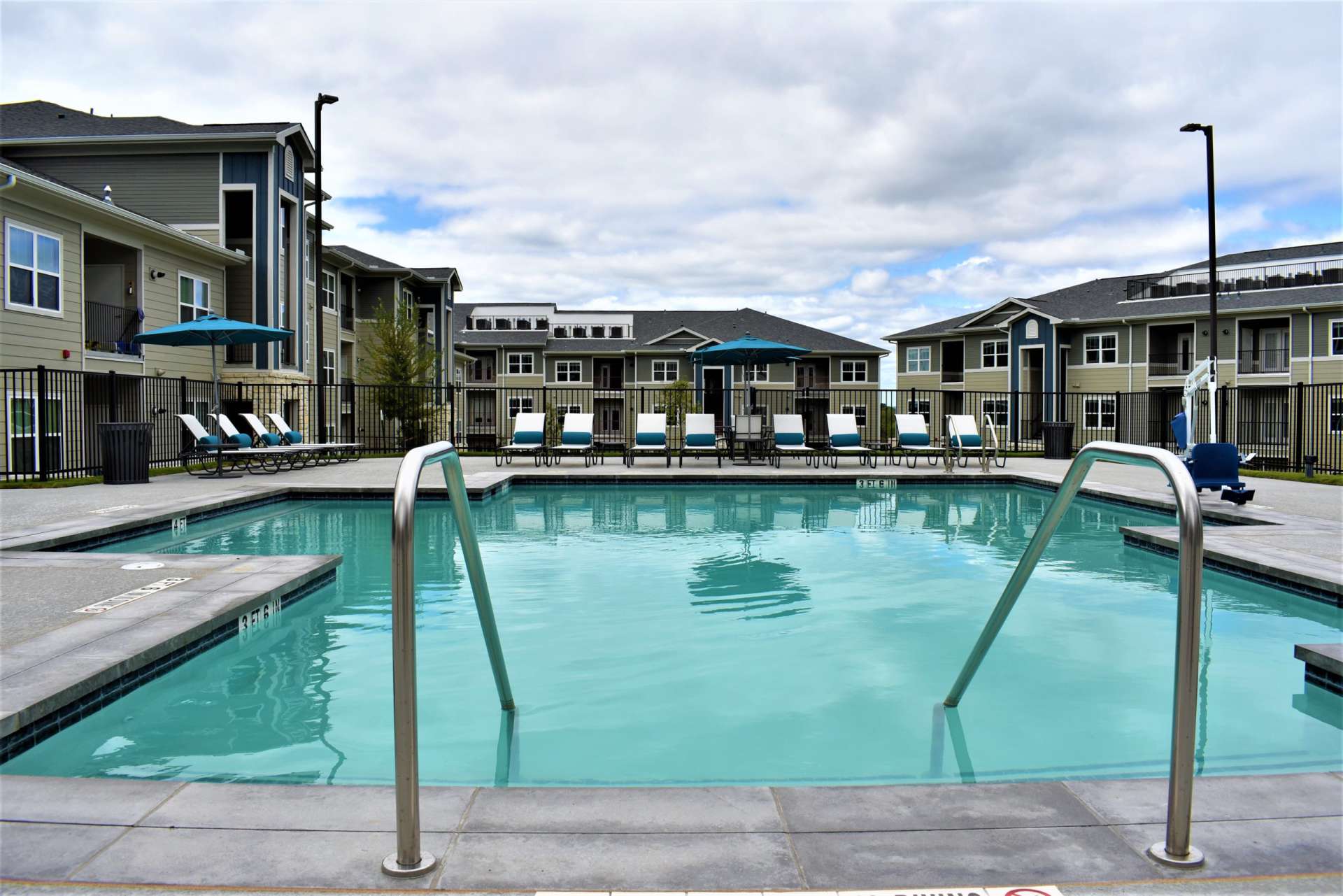 A view of a swimming pool from the deck area with the pool steps and handles in the foreground and lounge chairs, a chair lift for disabled individuals, and an apartment building in the background.